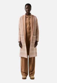 Long, open cardigan in a light peach knit with textured pattern and two front pockets, over a tan outfit and beige shoes.