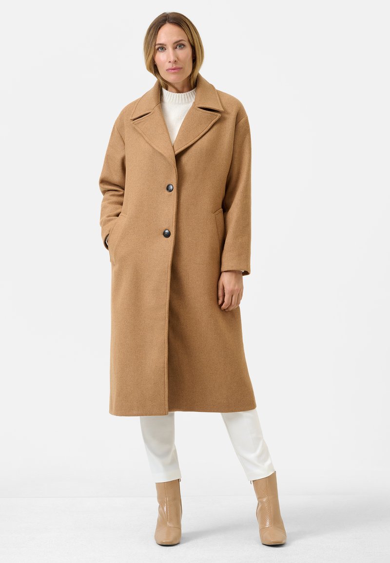 Bruine wollen jas met een geknipte kraag, twee voorzakken en twee donkere knopen. Draag het over een crèmekleurige trui en witte broek, gecombineerd met beige laarzen.