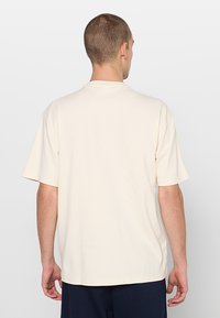 Lichtbeige T-shirt met korte mouwen en een ronde hals. Gladde textuur, losse pasvorm en minimalistisch ontwerp, gezien vanaf de achterkant.