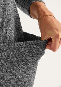 Grijze gebreide sweaterstof met een zachte textuur, met een zak die door een hand wordt vastgehouden. Het patroon toont ribbelstructuren en subtiele variaties.