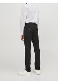 Černé slim-fit kalhoty s hladkou texturou, dvěma zadními kapsami a viditelnými knoflíky, kombinované s bílou košili s dlouhým rukávem a bílými teniskami.