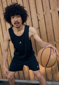 Débardeur sans manches noir et shorts avec un logo Jordan blanc, tenant un ballon de basket devant un fond en bois.