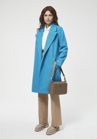Niebieski wełniany płaszcz z klapami, beżowe spodnie, beżowe mokasyny i szara torba na ramię z okrągłym emblematem. Gładka tekstura na całej powierzchni.