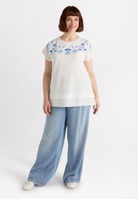 Top blanco con bordado floral azul en los hombros, combinado con jeans holgados de pierna ancha y color azul claro, y zapatillas blancas. Tejido suave.