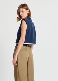 Gekürztes marineblaues ärmelloses Oberteil mit Kragen, weißer Besatz am Saum, kombiniert mit hoch taillierter Khaki-Hose mit weitem Bein. Glatte Textur.