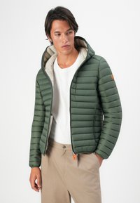 Bărbat îmbrăcat cu o jachetă căptușită verde cu glugă peste un tricou alb și pantaloni bej, stând cu o mână în buzunar și privind atent în lateral.