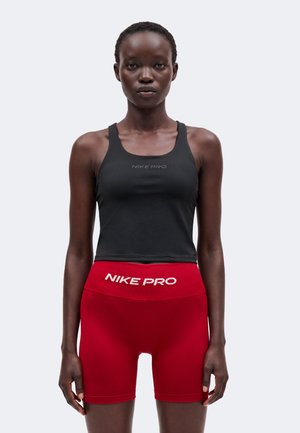 Femme portant un débardeur Nike Pro noir et un short de sport Nike Pro taille haute rouge sur un fond clair uni.