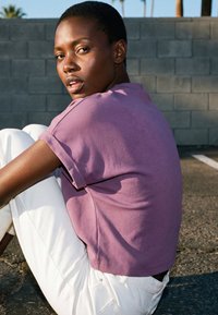 Mujer de cabello corto con camisa morada y pantalones blancos sentada sobre el asfalto, mirando por encima del hombro hacia la cámara.