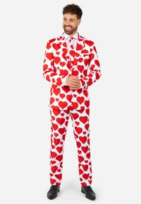 Biely oblek pokrytý veľkými červenými srdiečkami, so zodpovedajúcimi nohavicami a čiernymi formálnymi topánkami. Hladká látka, presne strihaný strih.