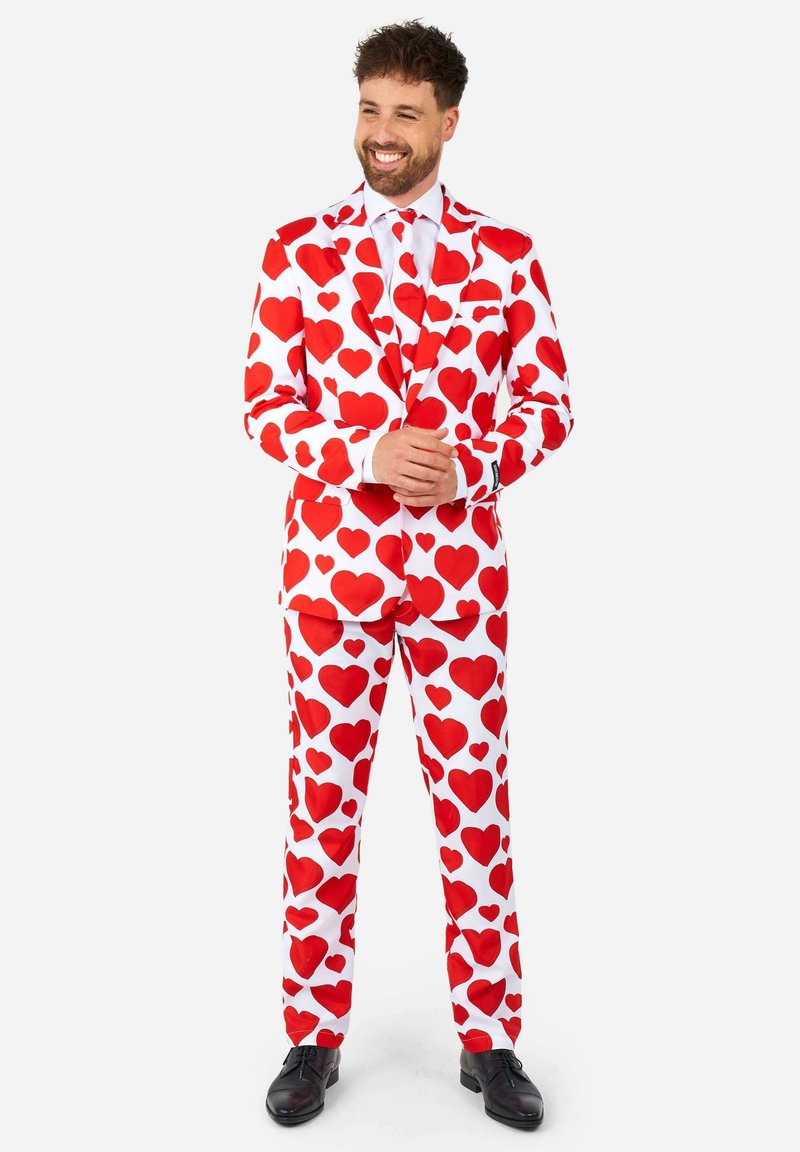 Biely oblek pokrytý veľkými červenými srdiečkami, so zodpovedajúcimi nohavicami a čiernymi formálnymi topánkami. Hladká látka, presne strihaný strih.