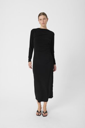 Schwarzes Langarmkleid mit Raffung, aus glattem Stoff gefertigt. Mit einem drapierten Ausschnitt und knielanger Länge. Trägt schwarze Flip-Flops.