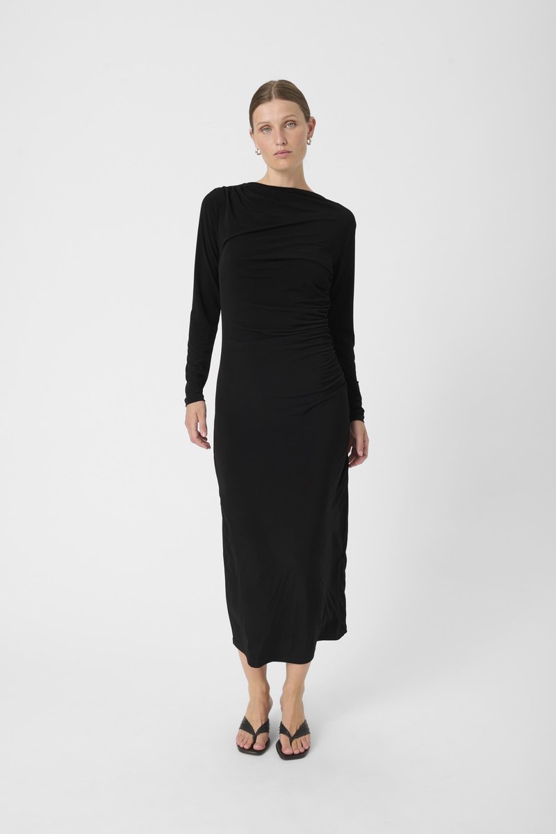 Schwarzes Langarmkleid mit Raffung, aus glattem Stoff gefertigt. Mit einem drapierten Ausschnitt und knielanger Länge. Trägt schwarze Flip-Flops.