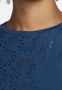 Camiseta deportiva azul con un patrón floral texturizado, mangas cortas y un logo negro de Nike en el pecho. Tela lisa con un ligero brillo.