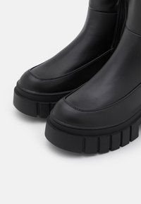 Bottines noires en matériau synthétique lisse, bout rond, semelle épaisse texturée et coutures visibles le long des coutures.