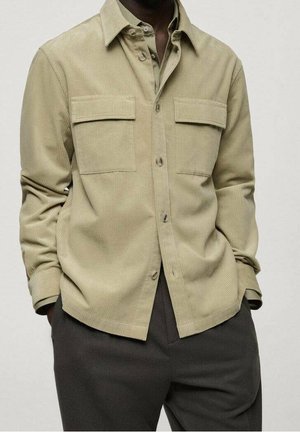 Mężczyzna ubrany w beżową koszulę sztruksową na guziki z klapkowymi kieszeniami, nałożoną na szarą koszulkę, w ciemnoszarych spodniach, z rękami w kieszeniach.