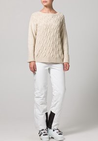 Beige gebreide trui met een kabelpatroon, gecombineerd met witte skibroek met zwarte accenten en een gestroomlijnde pasvorm.