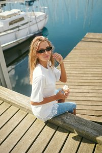 Femme avec des lunettes de soleil assise sur un quai en bois au bord de l'eau, tenant un cornet de glace, avec un bateau amarré à proximité par une journée ensoleillée.