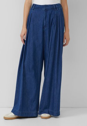 Wide-leg denim trousers in dark blue, featuring pleats at the waist, a straight cut, and a slightly ruffled texture, paired with beige sneakers.