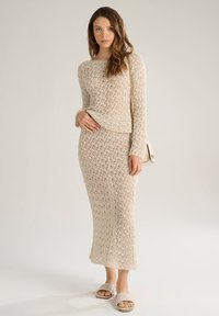 Set di maglia a due pezzi in beige chiaro, composto da un top a maniche lunghe e una gonna midi con motivo traforato; abbinato a scarpe slip-on beige.