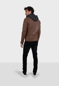 Giacca di pelle marrone con cappuccio grigio, design slim fit, tessuto morbido e cuciture visibili. Abbinata a pantaloni neri e sneaker bianche.