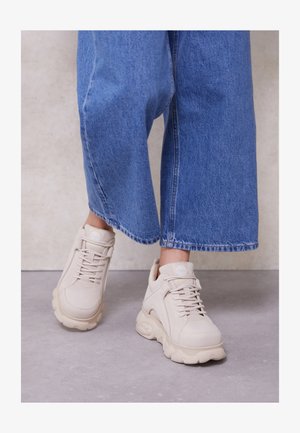 Világos bézs sportcipő sima textúrával, lekerekített orral, vastag talppal és fűzőkkel. Széles szárú kék farmernadrággal párosítva.