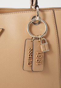 Handtas met een structuur in naturel kleur, voorzien van een gouden ring, twee labels met reliëf "GUESS" en "1981", en een kleine bijpassende hangslot bevestigd aan het handvat.