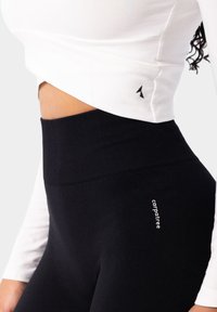 Balta kultūra augšdaļa ar sānu logotipu un melniem augsto vidukļa legingiem ar drukātu zīmola nosaukumu. Materiāls ir gluds ar nelielu stiepšanos.