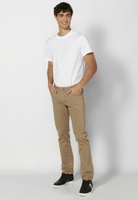 Pantalones de algodón beige ajustados, combinados con una camiseta blanca de manga corta y zapatillas negras. Con bolsillos frontales y botón de cierre.