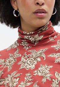 Gola alta vermelha com padrão de flores brancas e marrons, textura suave e pescoço ajustado e canelado. Brincos de argola prateados visíveis.