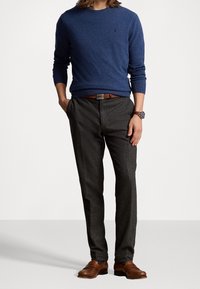 Homme portant un pull bleu, un pantalon gris foncé, une ceinture en cuir marron, une montre et des mocassins en cuir marron, debout avec les mains partiellement dans les poches.