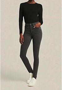 Jean skinny noir taille haute avec une texture lisse et un léger éclat, associé à un haut ajusté à manches longues et des baskets blanches.