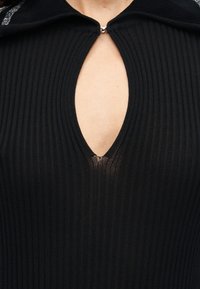 Κοντινό πλάνο μαύρου πλεκτού τοπ με ριμπ ύφανση, μικρό ασημένιο κούμπωμα στο λαιμό και άνοιγμα σε σχήμα κλειδαρότρυπας που αφήνει ακάλυπτο το δέρμα.