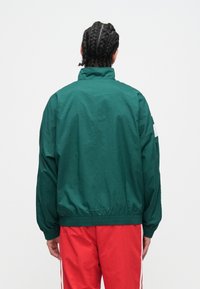 Osoba s zapletenými vlasmi, oblečená v tmavozelenej bunde a červených nohaviciach, stojaca čelom k jednoliatemu bielemu pozadiu.