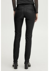 Pantalons skinny en simili cuir noir avec une taille haute, des coutures discrètes, des poches à l'arrière et une texture lisse. Portés avec des chaussures à talons noires.
