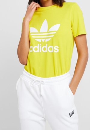 Gul bomuld T-shirt med hvidt Adidas-logo og tre-stribet design, kombineret med hvide joggingbukser med snøre og sort mærke.