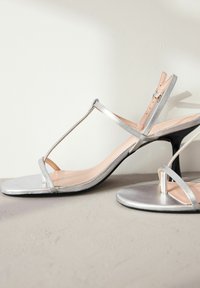 Zilveren metallic hoge hakken sandalen met een T-band ontwerp, open teen en een gladde textuur. Bevat een zwarte hak en een naakte binnenzool.