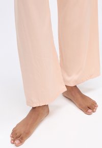 Losse, lichtperzikkleurige broek met een gladde textuur en wijde pijpen, met een schone, rechte zoom. Blote voeten met lichtroze nagellak zichtbaar.