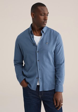 Uomo che indossa una camicia blu abbottonata sopra una t-shirt bianca e jeans scuri, con una mano in tasca, che guarda di lato.