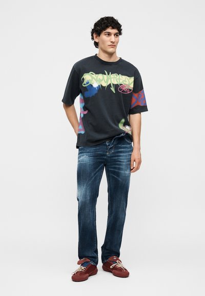 Νέος άνδρας που στέκεται φορώντας μαύρο κοντομάνικο με στάμπα graffiti, ξεθωριασμένα μπλε τζιν και κόκκινα αθλητικά παπούτσια σε ένα απλό φωτεινό φόντο.