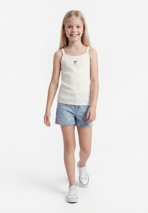 Šypsosi jauna mergina vaikšto, dėvi kreminės spalvos tanką su mažu kaspinėlio raštu, šviesiai mėlynas džinsines trumpikes ir baltus sportinius batelius.