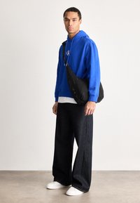 Egy férfi élénk kék kapucnis pulóverben, széles fekete nadrágban, fehér tornacipőben áll, és egy fekete keresztpántos táska van nála, egy sima háttér előtt.