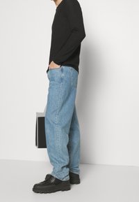 Person in hellblauen, lässig geschnittenen Jeans, schwarzem Langarmshirt und schwarzen klobigen Schuhen, der vor einem weißen Hintergrund steht.