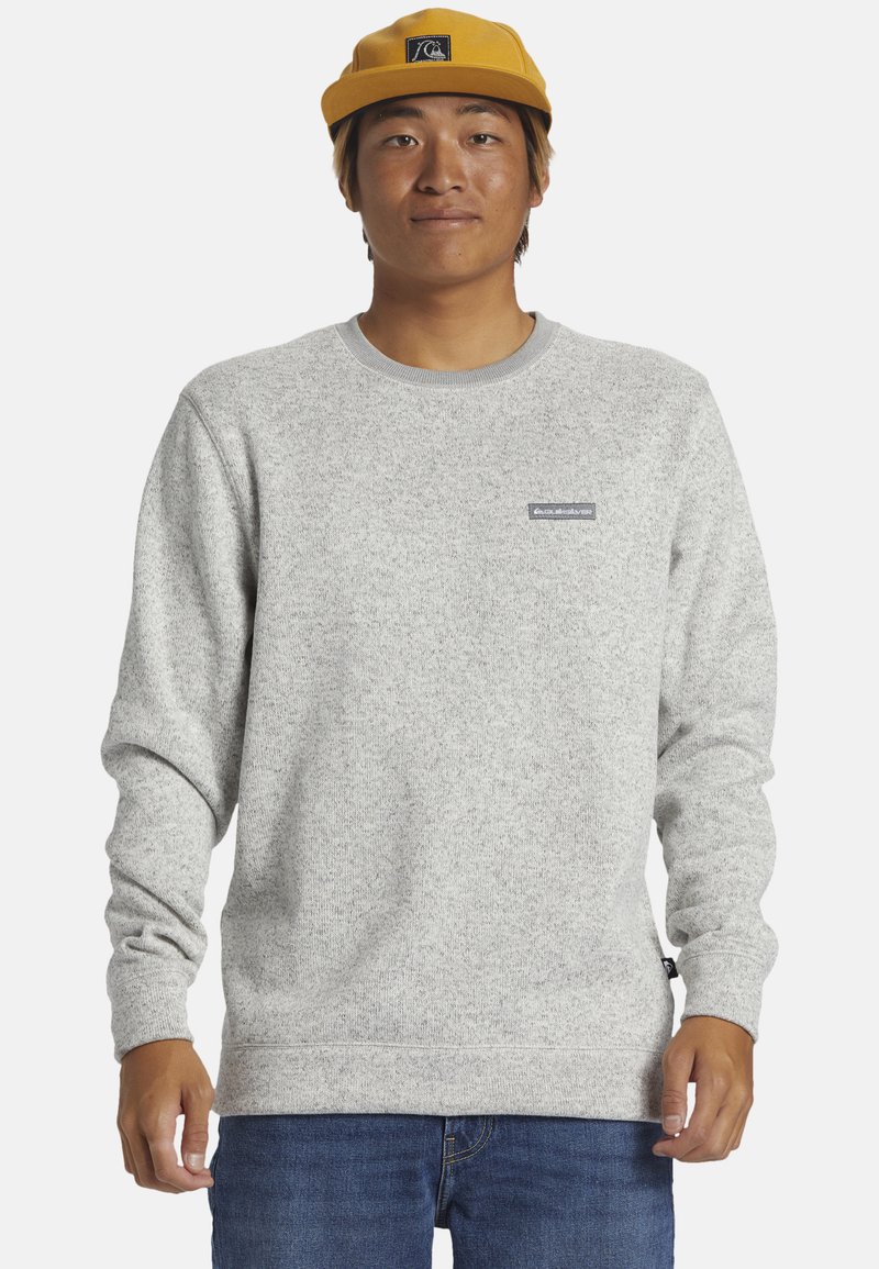 Helle graue Sweatshirt aus strukturiertem Stoff, mit Rundhalsausschnitt und gesticktem Logopatch auf der Brust. Kombiniert mit blauen Jeans.