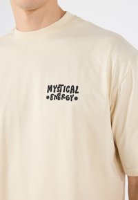Beige t-skjorte laget av bomull, med et svart grafisk trykk som sier "MYSTICAL ENERGY" på venstre side av brystet.