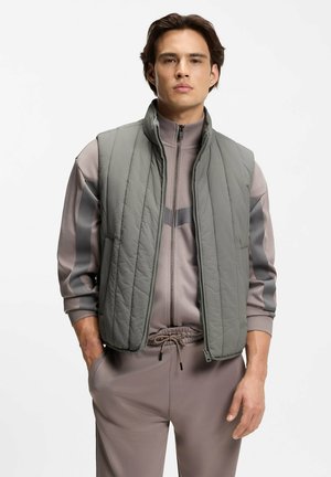 Mann trägt graue gesteppte Weste über einem mauve-grauen Reißverschluss-Sweatshirt und passenden Hosen, steht mit den Händen in den Taschen vor weißem Hintergrund.