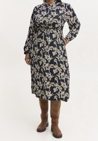 Lång marinblå klänning med blommönster, krage, smockad midja, långa ärmar och fickor. Bärs med bruna knähöga stövlar.