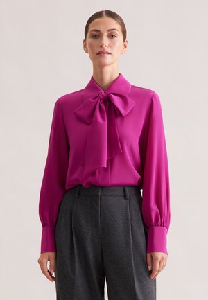 Blusa fucsia con un grande fiocco al colletto, maniche lunghe con polsini aderenti, abbinata a pantaloni grigi a vita alta. Tessuto dalla texture liscia.