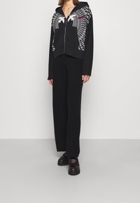 Haut à fermeture éclair noir avec un design graphique blanc et des lettres sur le devant. Accompagné de pantalons noirs et de chaussures à plateforme noires.