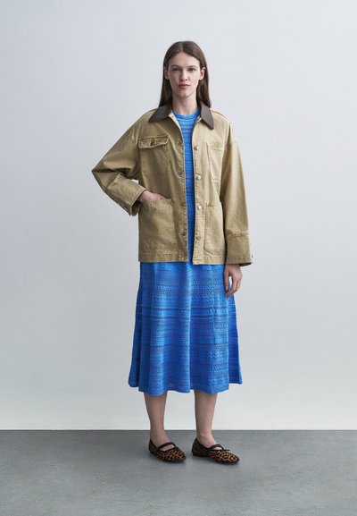 Une femme debout porte une veste surdimensionnée de couleur beige avec de grandes poches, une robe midi bleue à motifs, et des chaussures Mary Jane imprimé léopard, sur un fond uni.