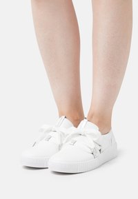 Sneaker bianchi con suola in gomma testurizzata, ampie stringhe bianche e dettagli in metallo argentato, caratterizzati da una finitura liscia e una punta arrotondata.